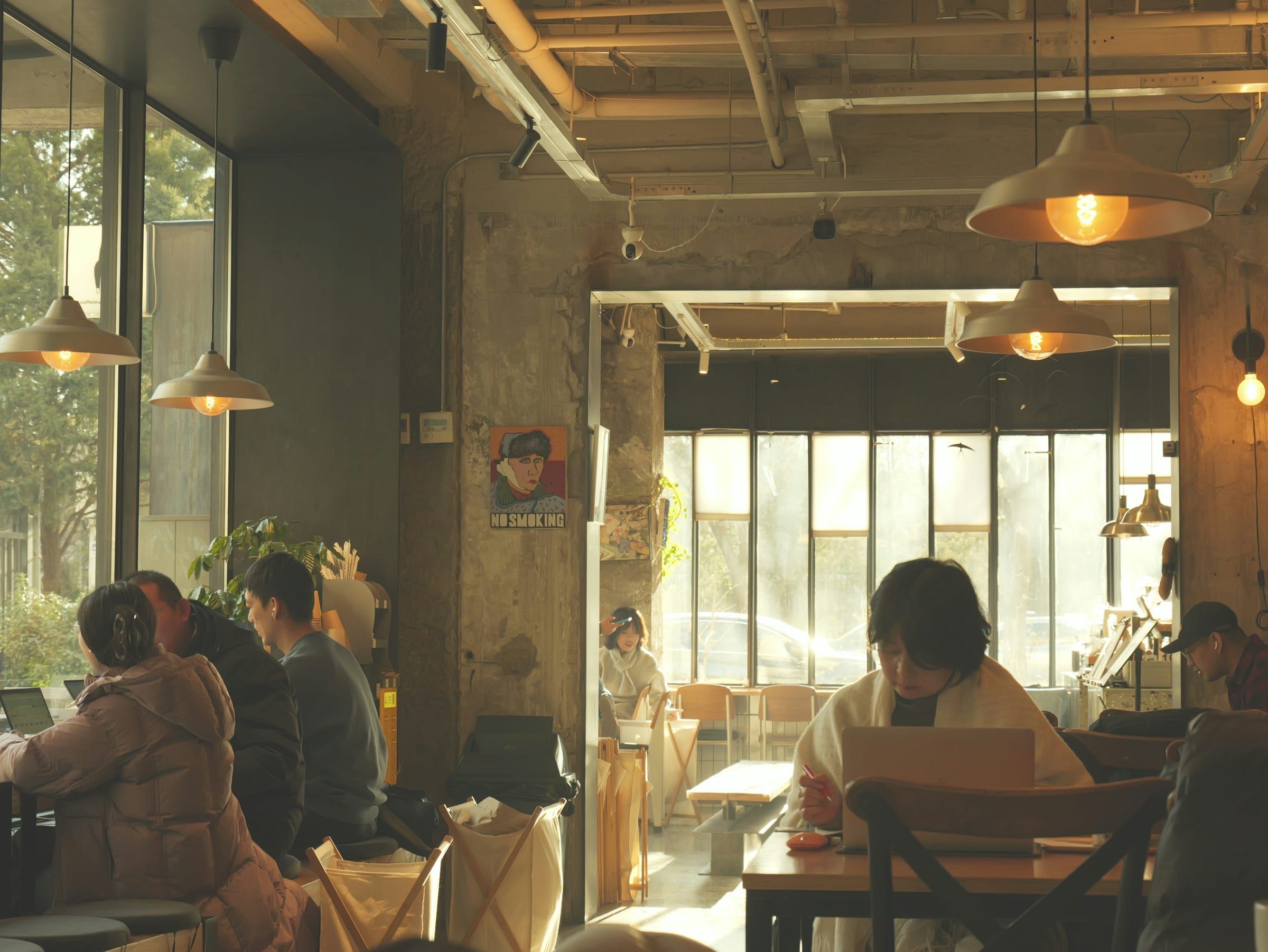 A sunlit cafe with tall windows and pendant lamps, where people work and talk at wooden tables