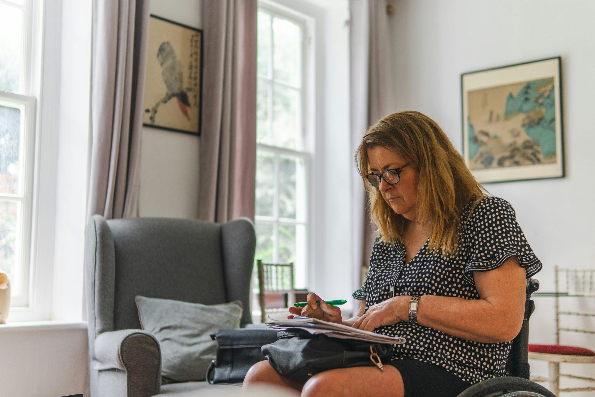 A woman in glasses reads through papers in her lap in a bright living room with an armchair and framed prints