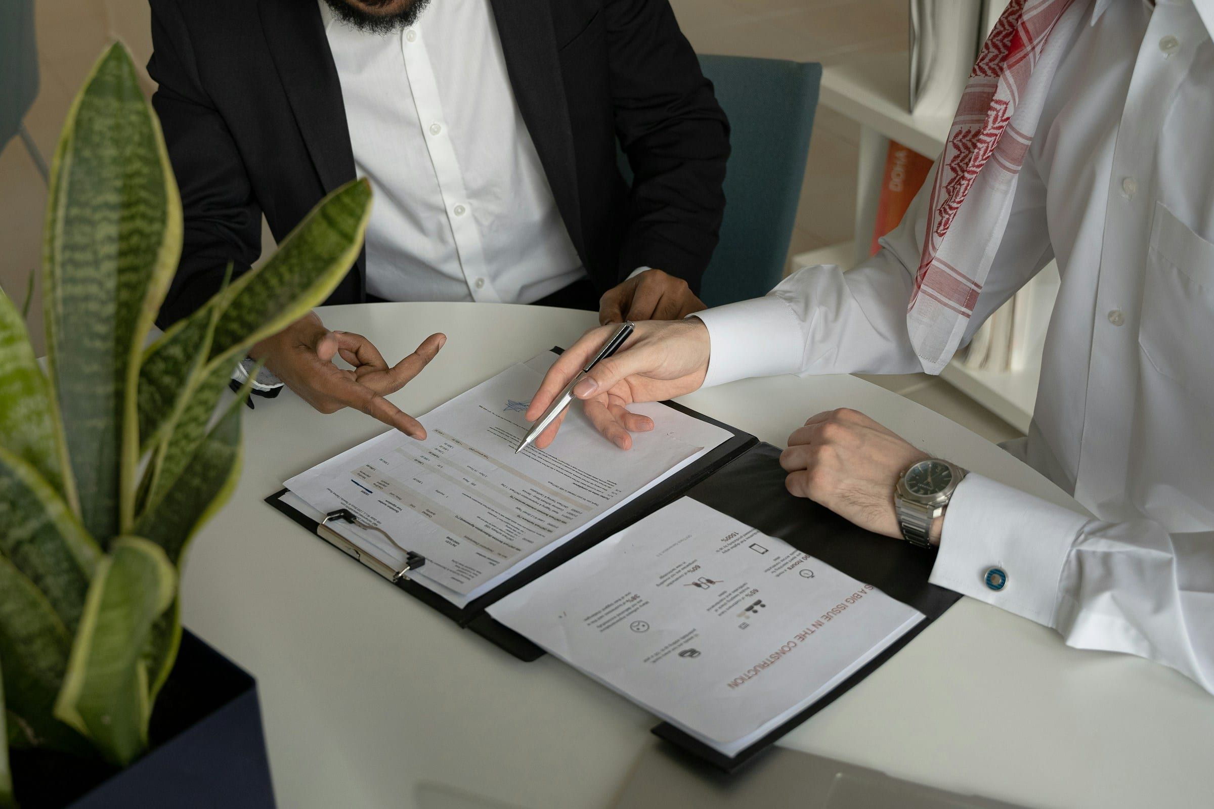 Two men review documents together at a white table, one in a dark suit and one in a white thobe and red-and-white keffiyeh