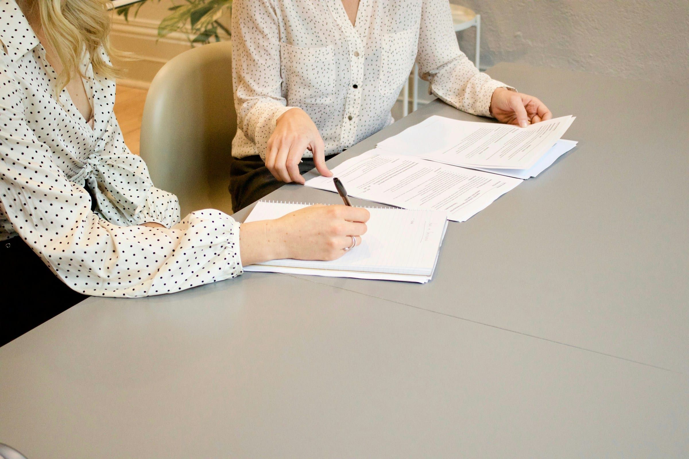 Two women sit at a pale table, one signing a printed document while the other reads through the pages