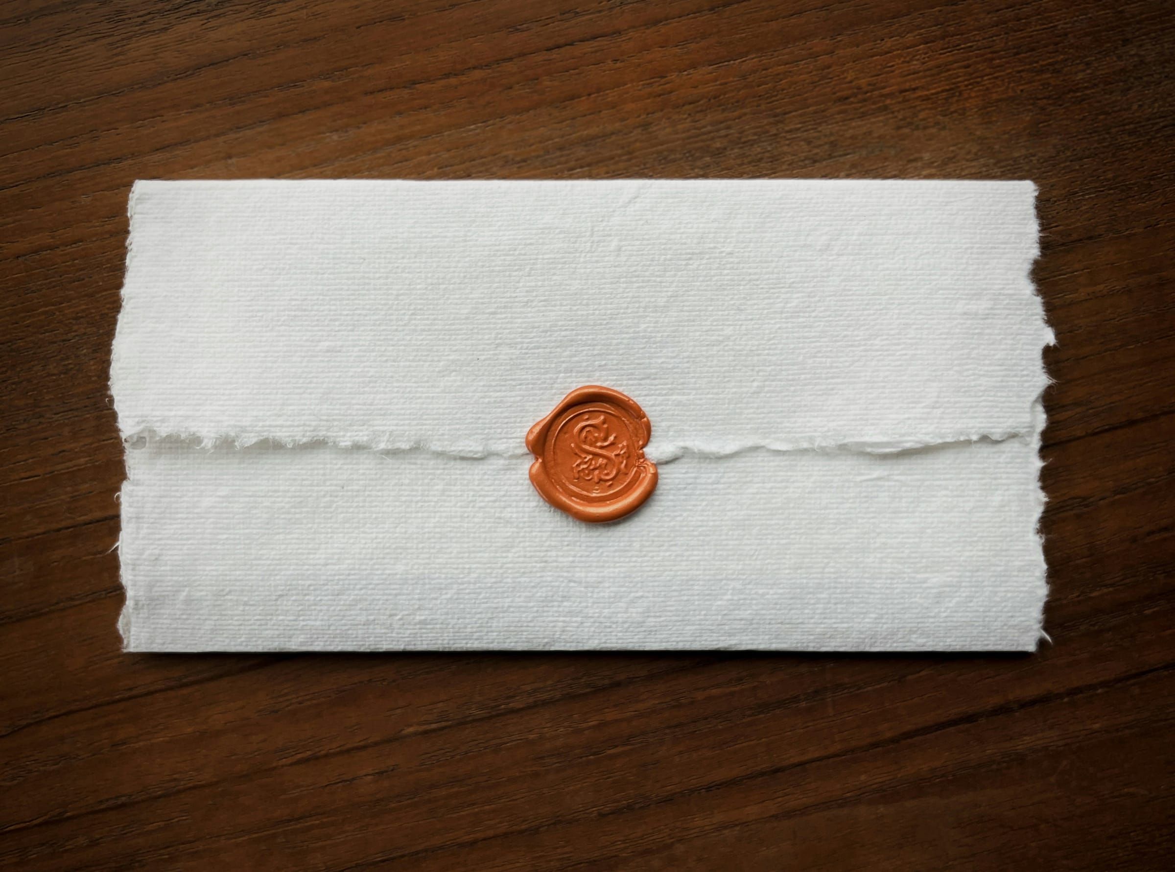 Um envelope de papel branco com bordas irregulares lacrado com um selo de cera laranja sobre uma mesa de madeira escura