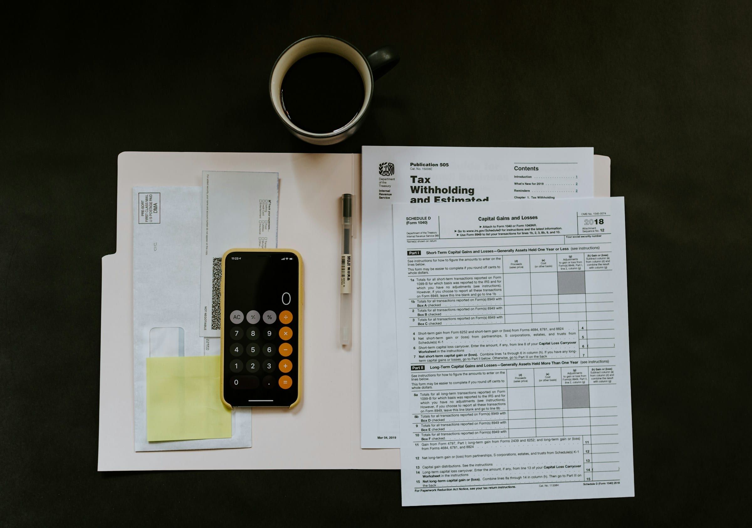 Top-down view of blank US tax forms beside a phone calculator, a pen and a cup of black coffee on a dark desk