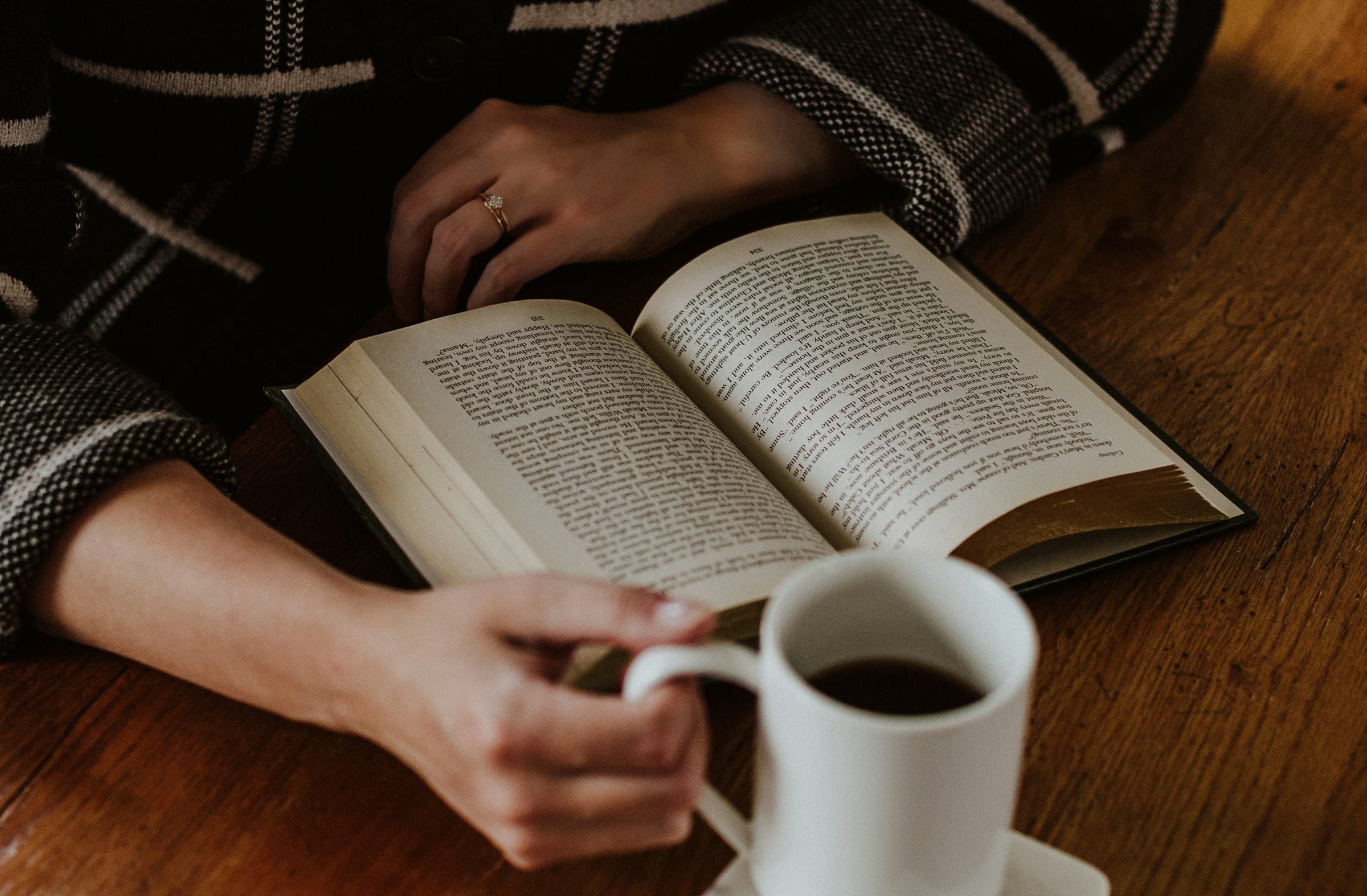 Primo piano di una persona che legge un libro aperto su un tavolo di legno tenendo una tazza di caffè nero