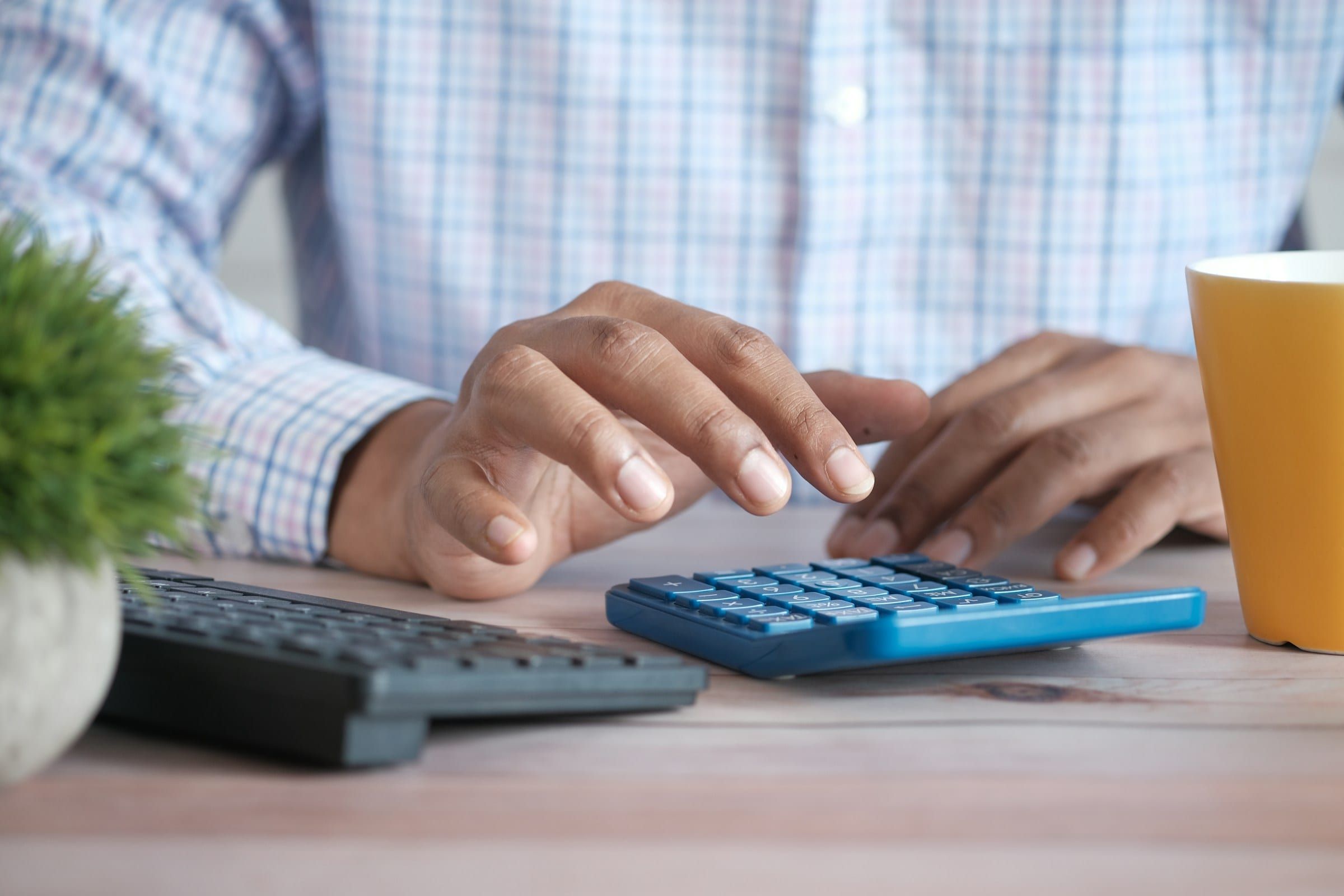 A man in a checked shirt works a blue calculator beside a keyboard and a yellow mug