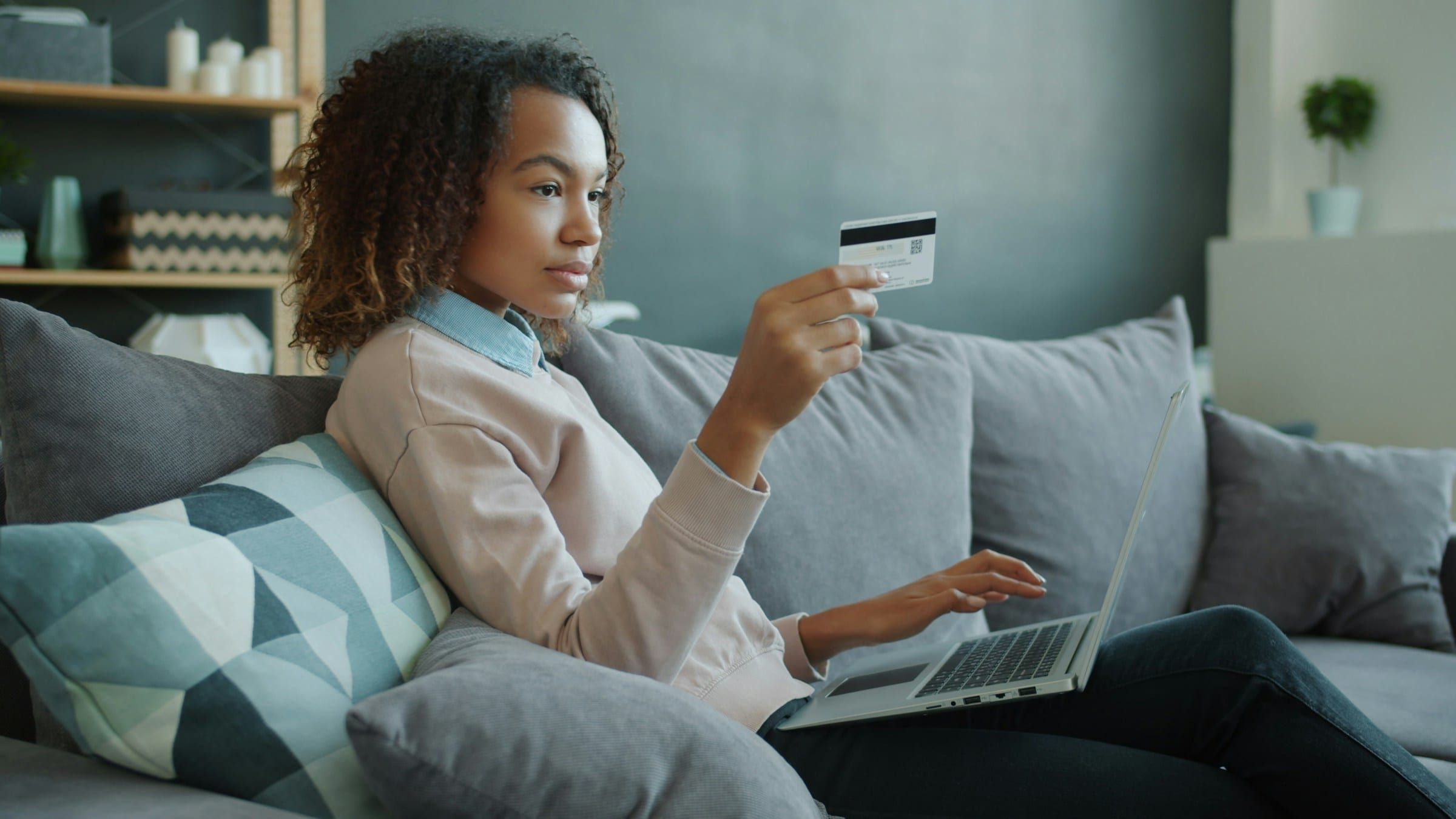 Una donna su un divano grigio mostra una carta bancaria mentre usa un laptop sulle ginocchia