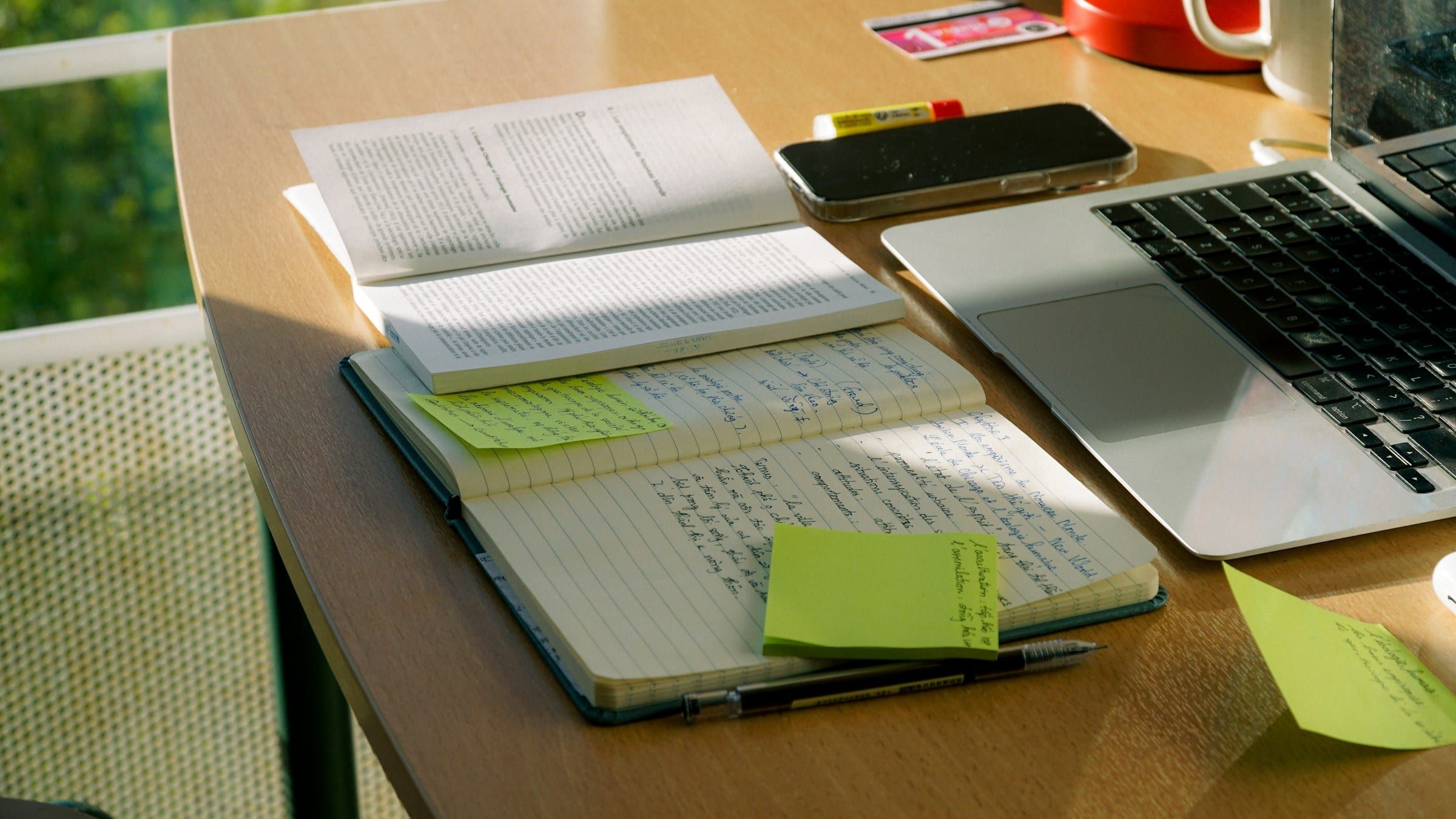 Un quaderno aperto con appunti scritti a mano e post-it verdi accanto a un laptop su una scrivania soleggiata