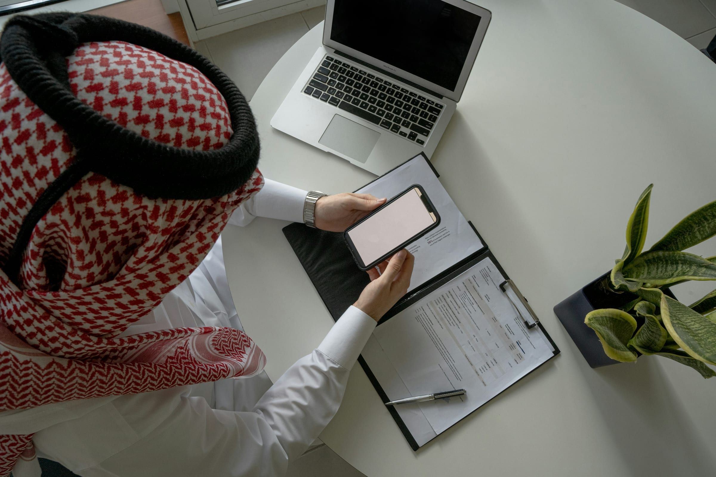 Top-down view of a man in a red and white keffiyeh holding his phone over a clipboard, next to a laptop and a plant