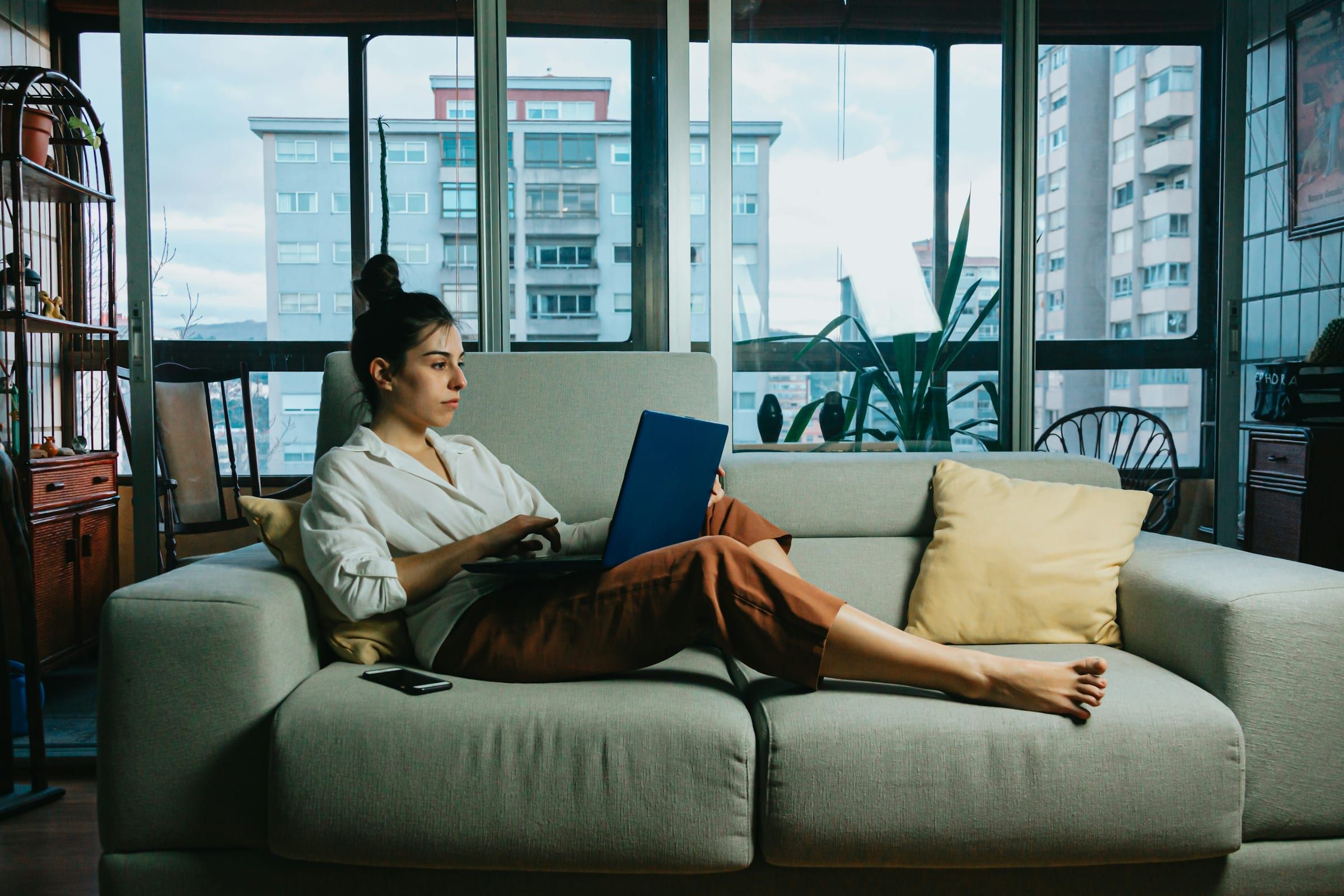A woman works on a laptop while stretched out on a sage-green sofa beside tall city windows
