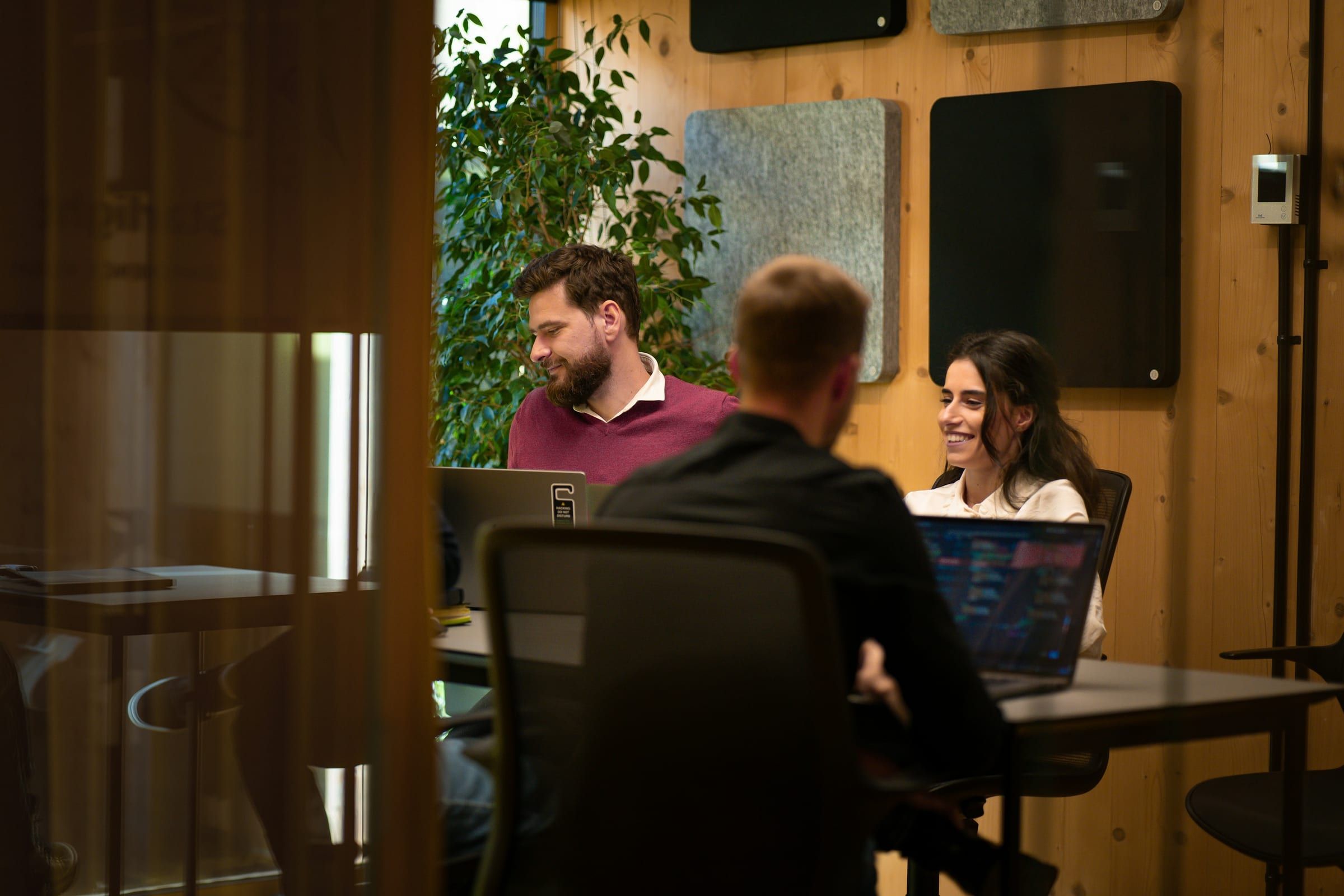 Three colleagues talk and smile around a table of laptops in a meeting room with warm wooden walls and a tall plant