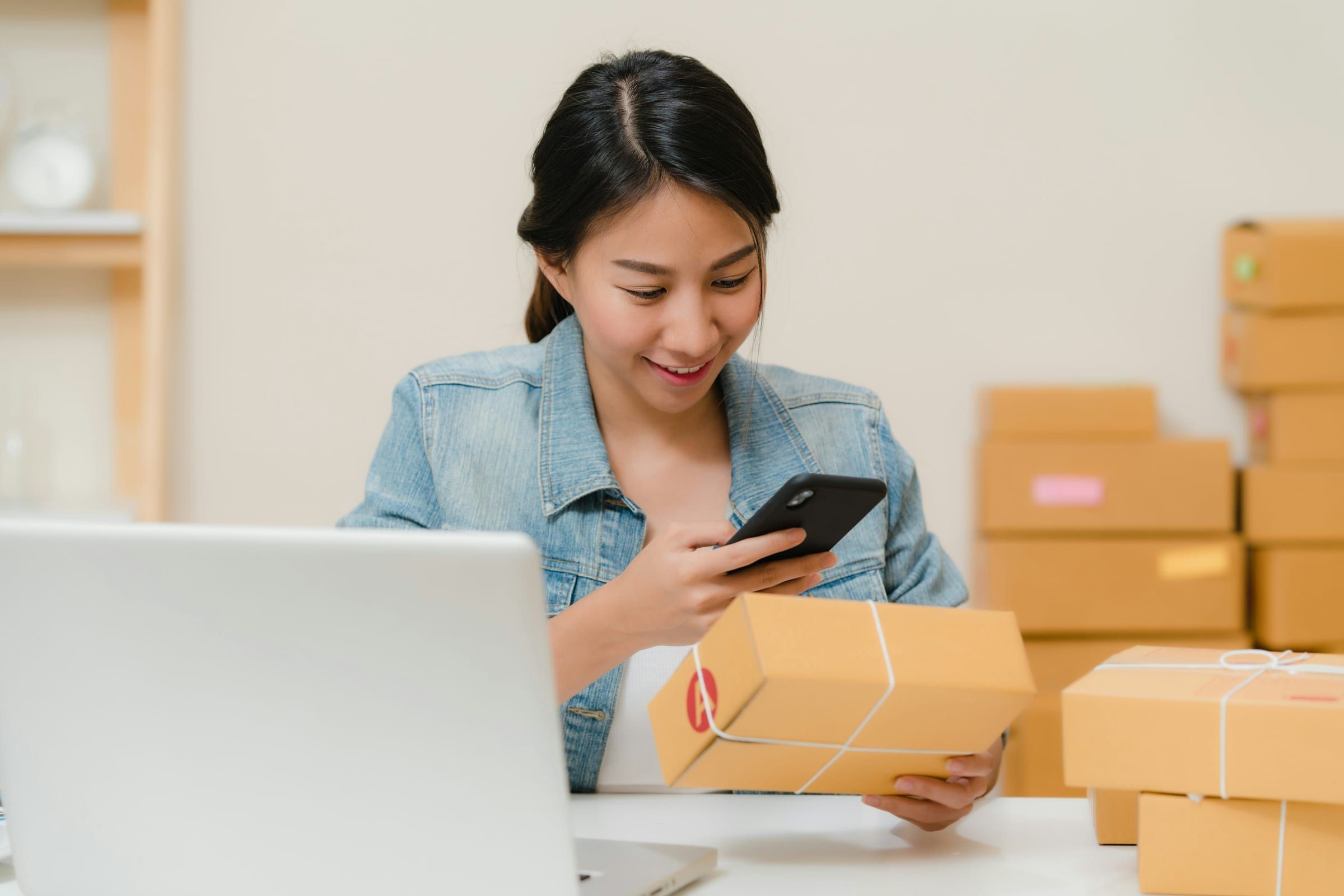 A smiling woman checks a parcel against her phone at a desk surrounded by stacked shipping boxes