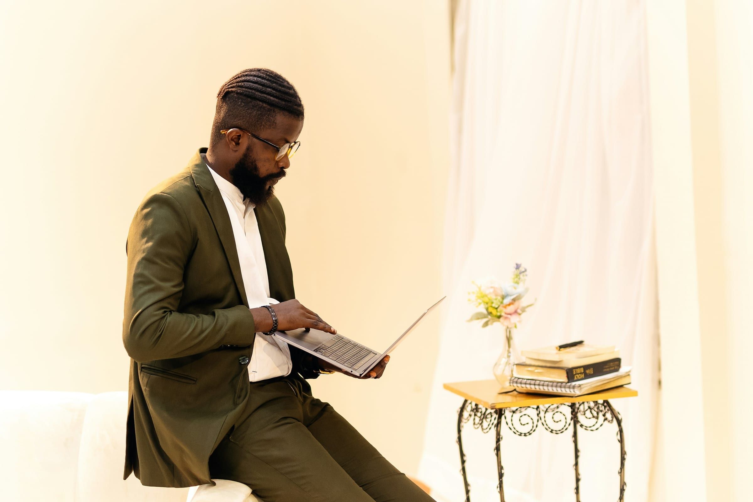 A man in an olive suit sits on a white chair reading from an open laptop in a softly lit cream room