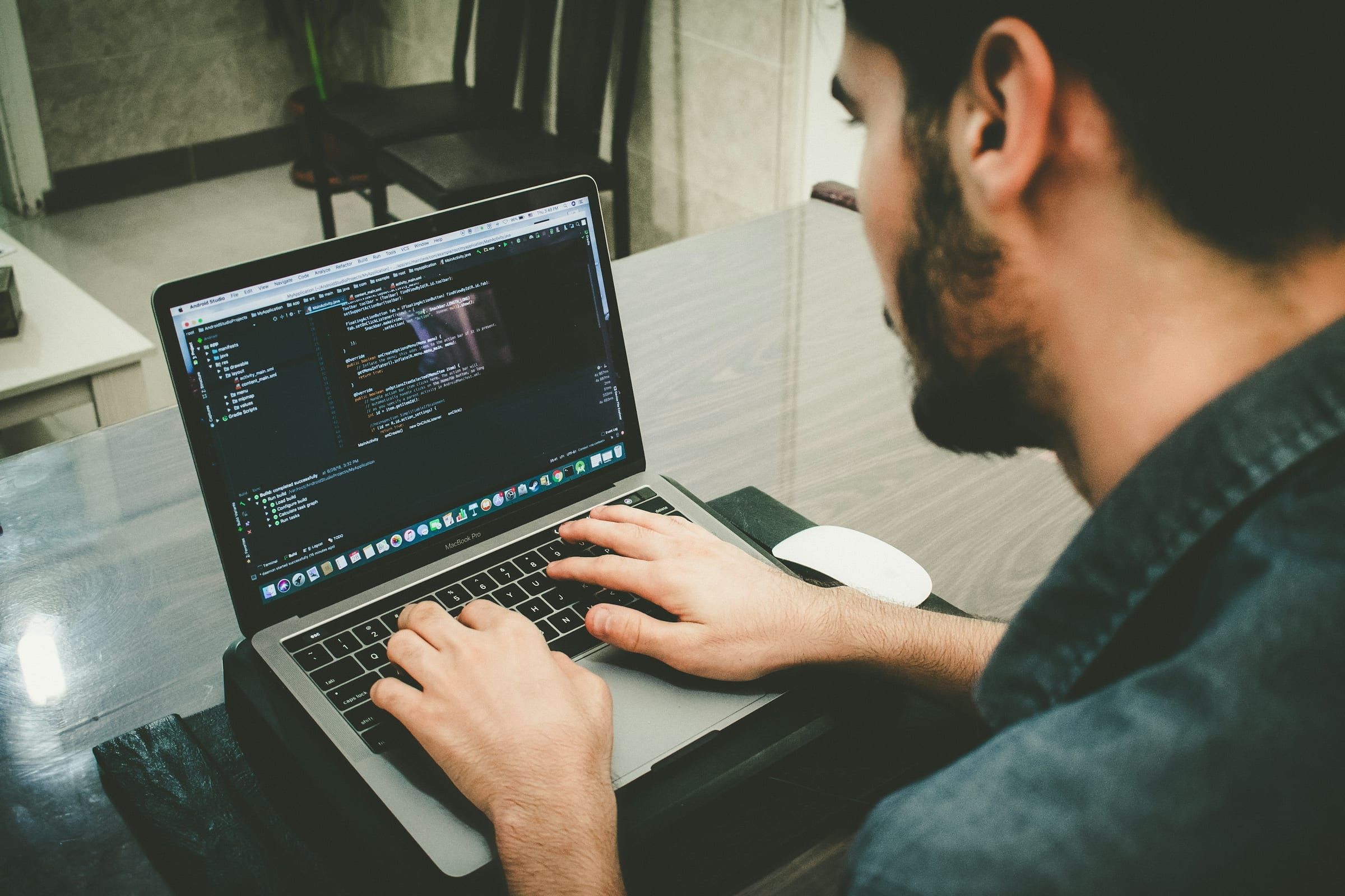 Over the shoulder of a bearded man writing code on a laptop at a glass table