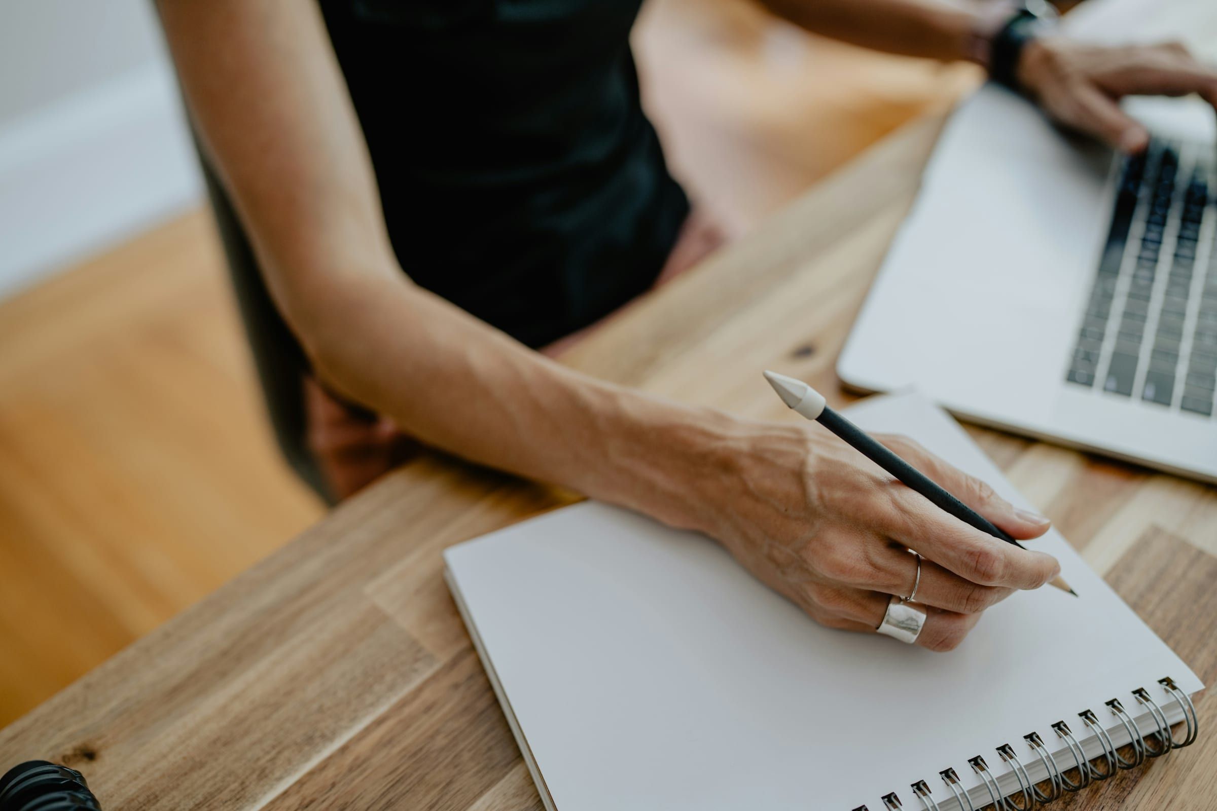 Close-up of a hand writing with a pencil in a blank spiral notebook next to an open laptop on a wooden desk