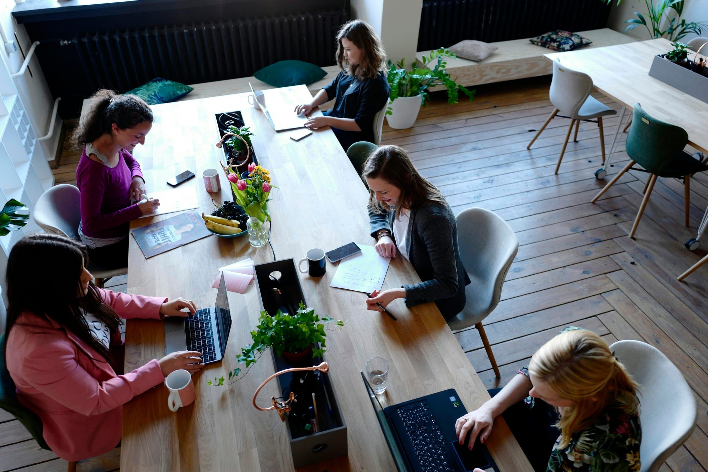 Seen from above, five women work on laptops and papers around a long wooden table with flowers in a coworking space