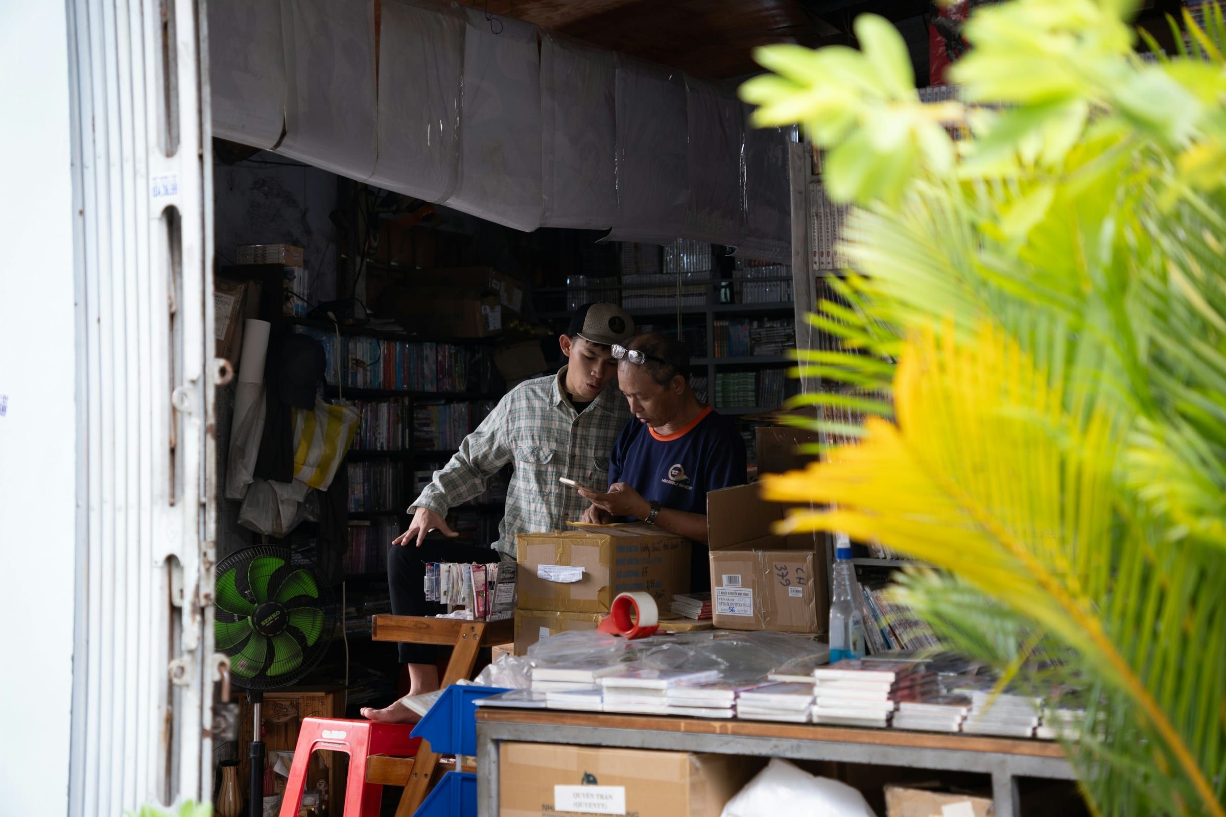 Two people sort stock and parcels at a long counter in a small, crowded shop, framed by a green plant