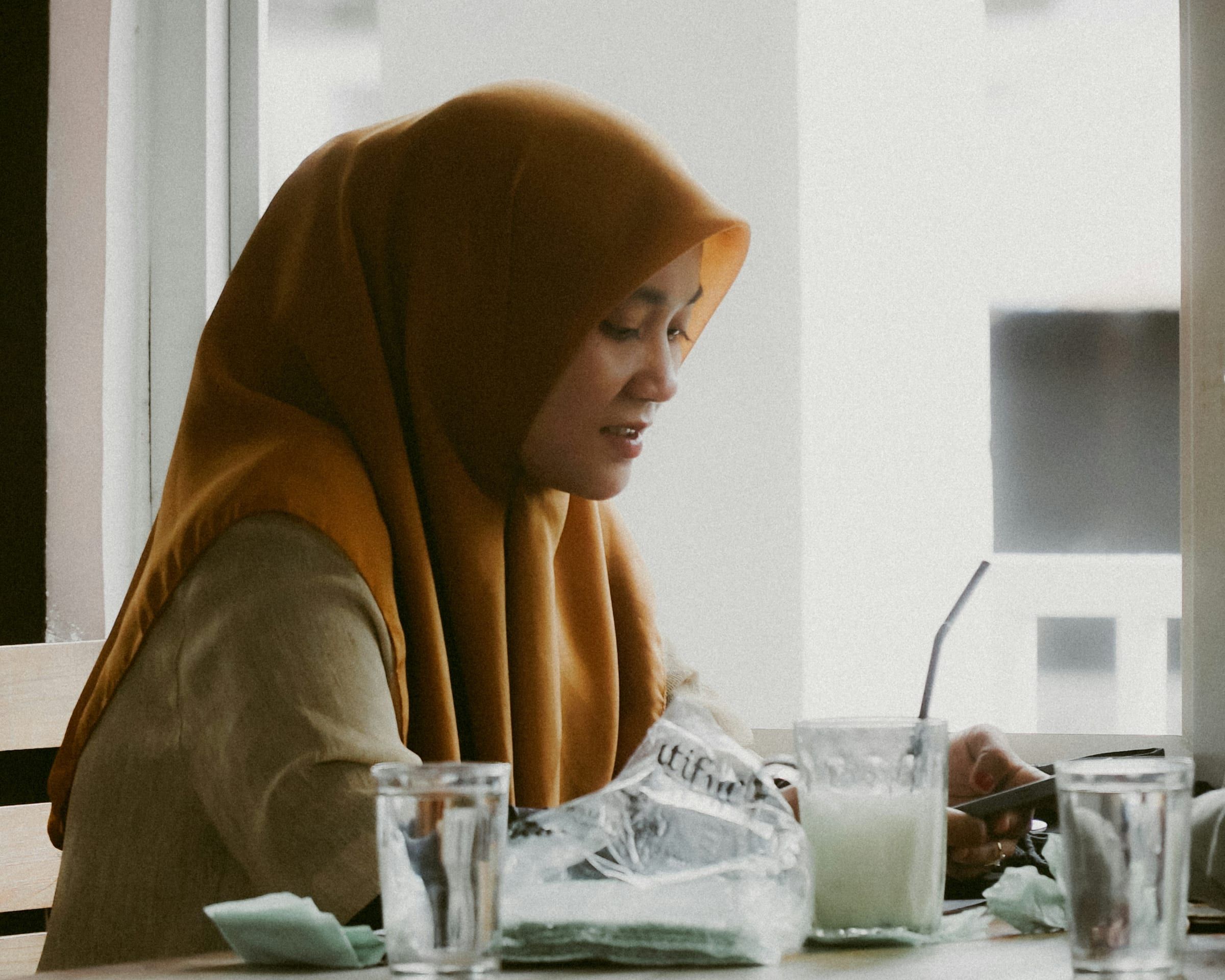 A woman in an ochre hijab looks down at her phone at a cafe table with glasses of water and a drink