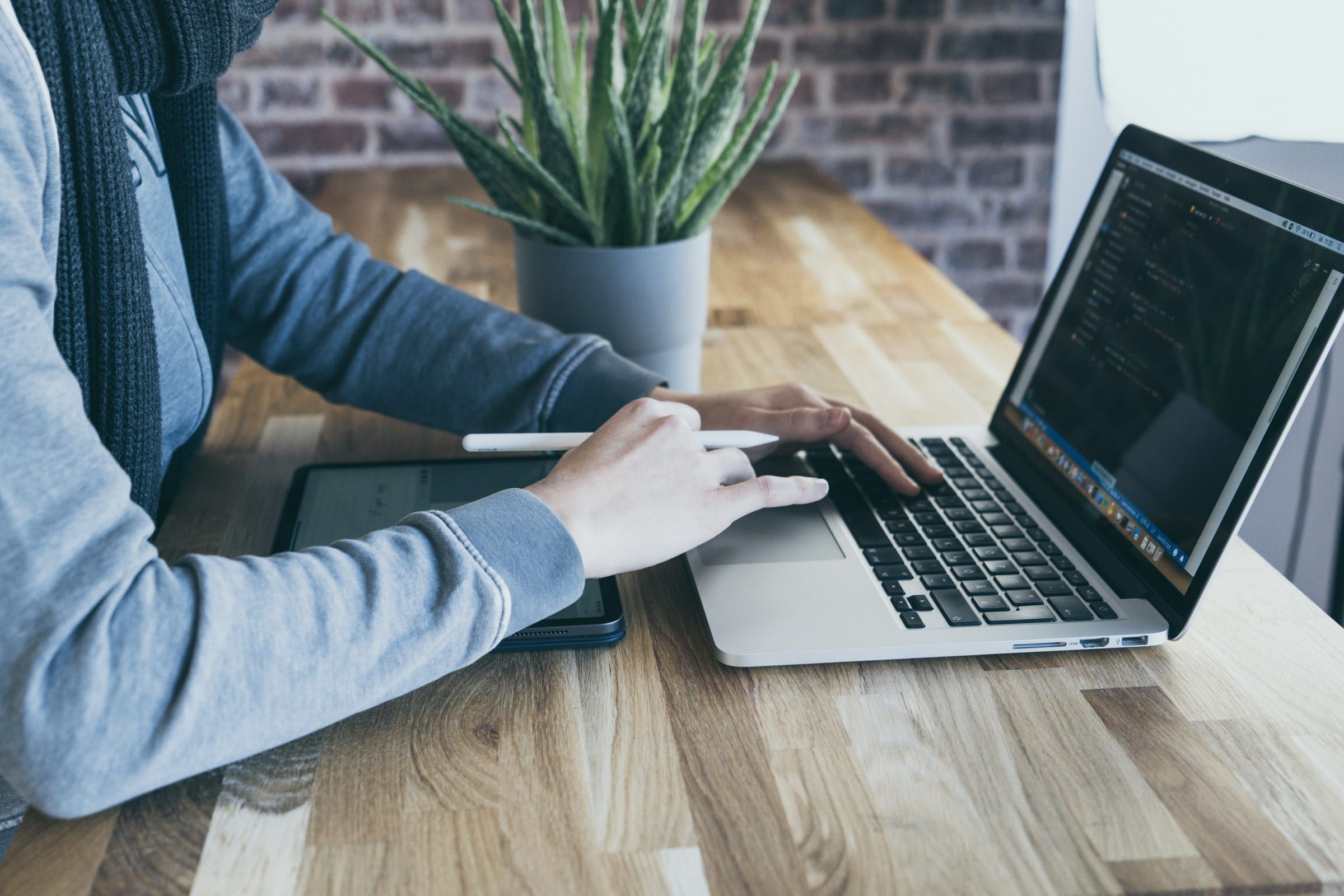 Hands typing on a laptop with a dark code editor, next to a tablet and a potted plant on a wooden table